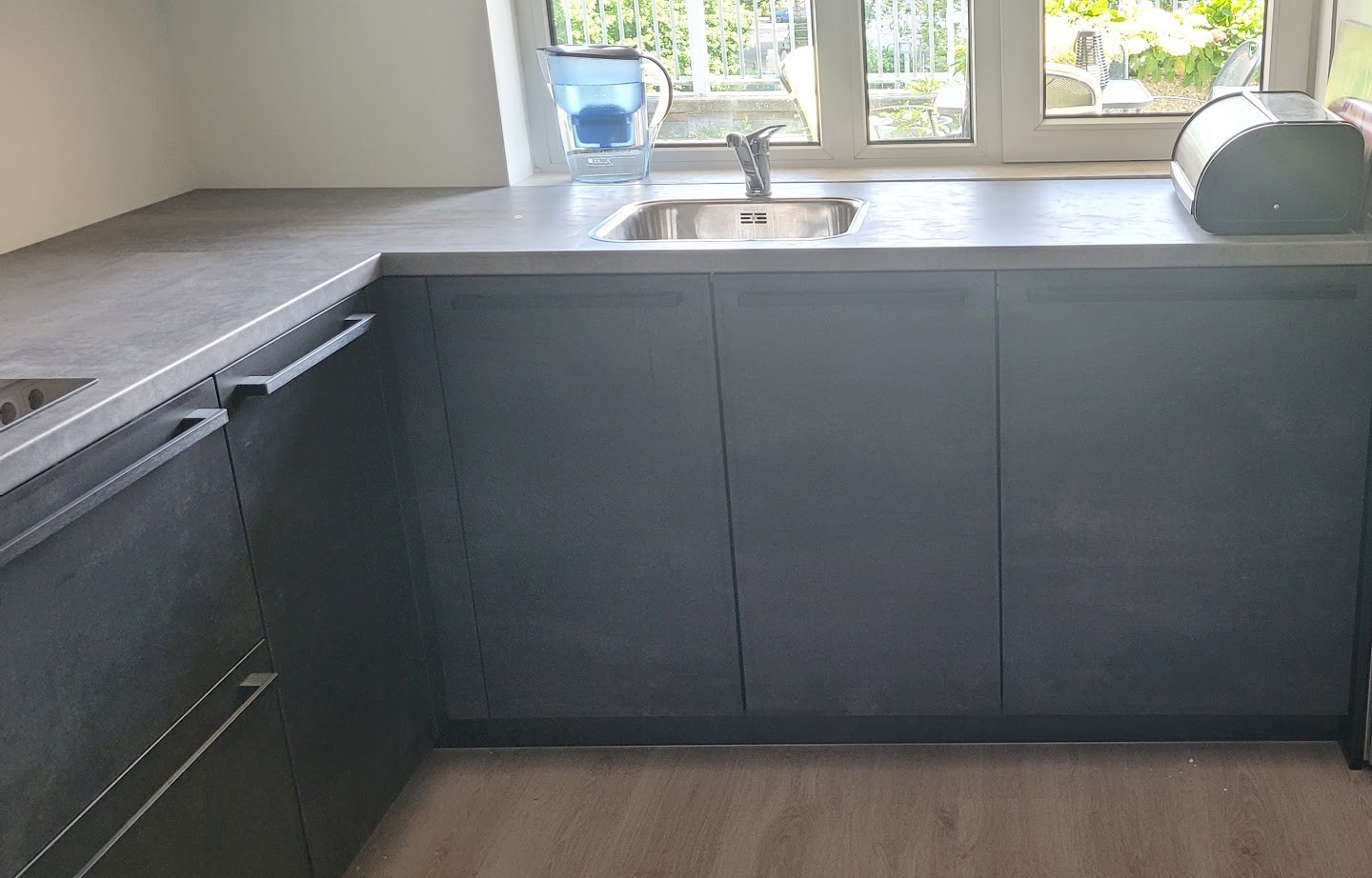 Antraciet spoelkeuken onder het raam met betonlook werkblad en een rvs koelkast